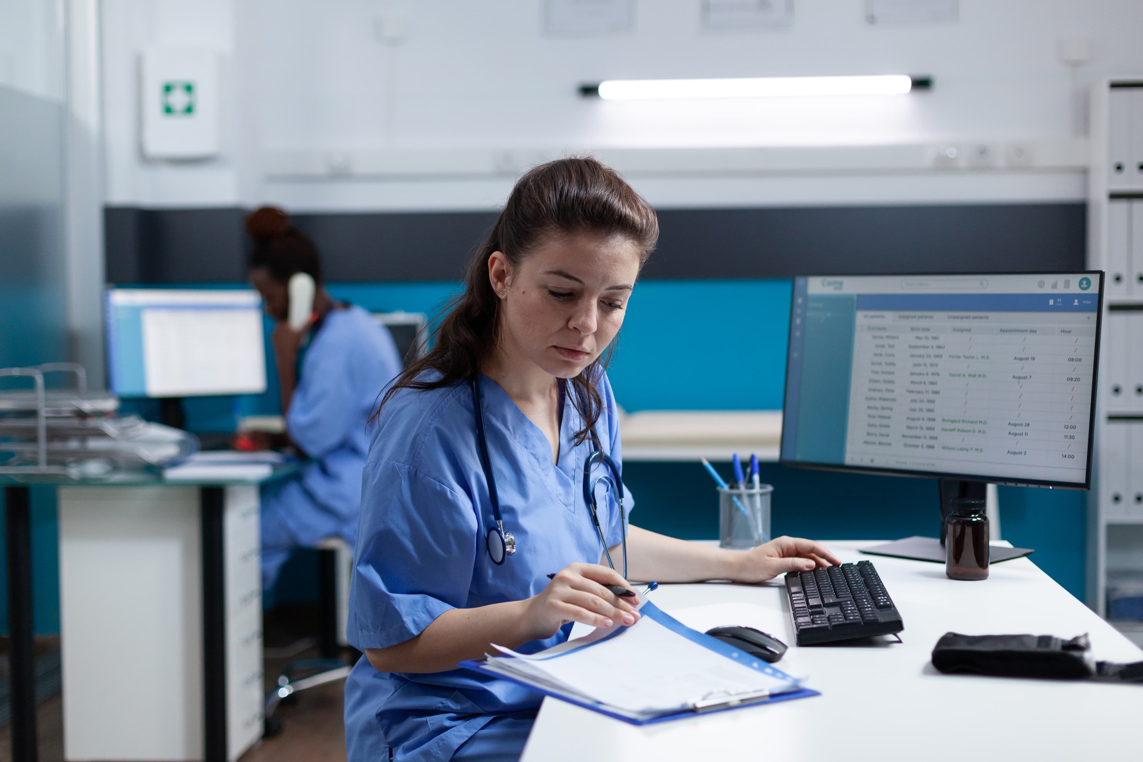 Care manager in scrubs reviewing a patient chart at a clinical workstation, with a colleague on a phone call in the background